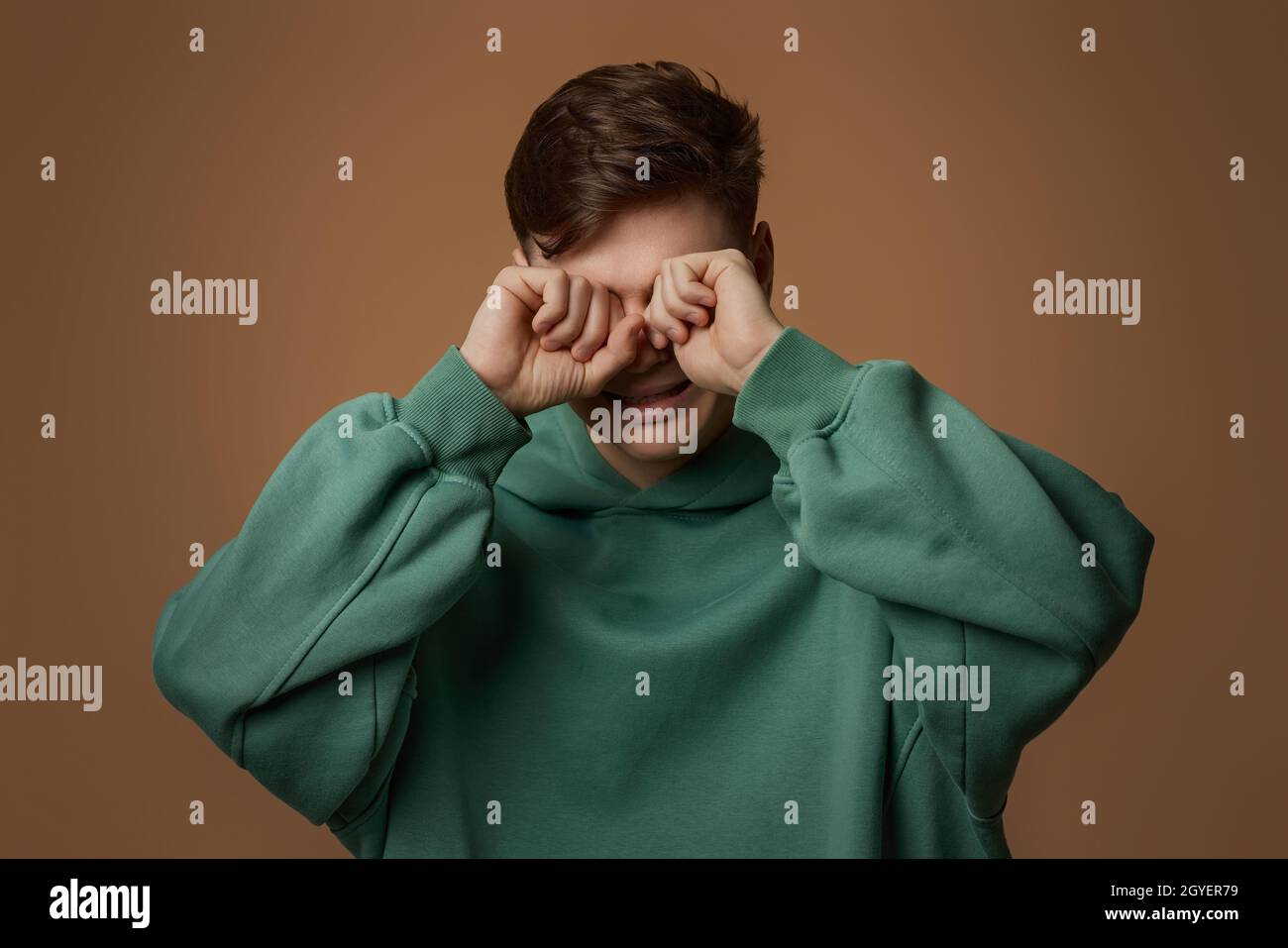 Verärgert weinender junger Mann im Sweatshirt isoliert auf braunem Hintergrund. Stockfoto