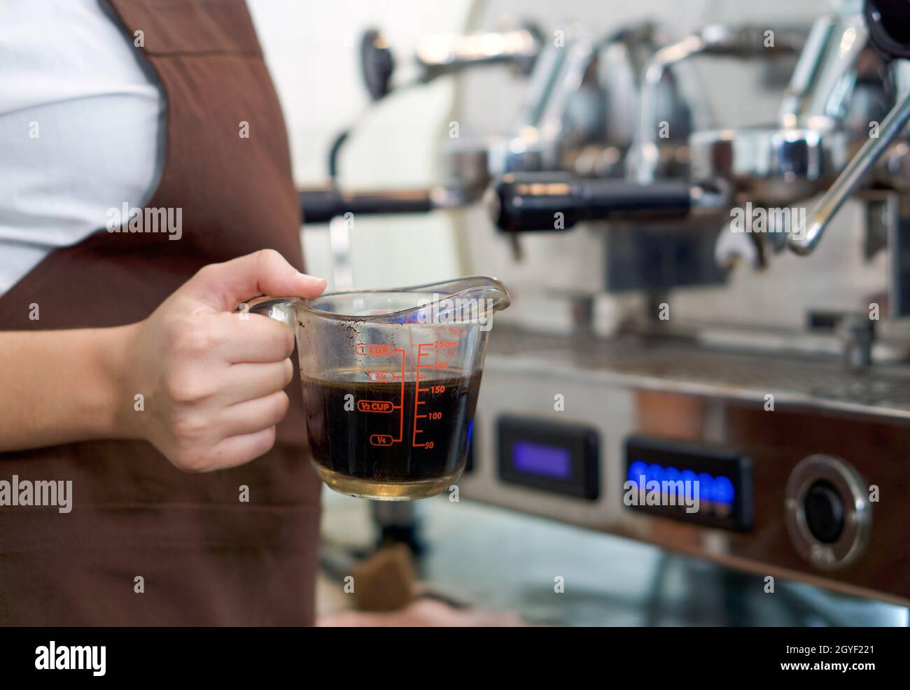 Barista in brauner Schürze mit einer Messbecher gefüllt mit heißem Kaffee. Morgendliche Atmosphäre in einem Café. Stockfoto