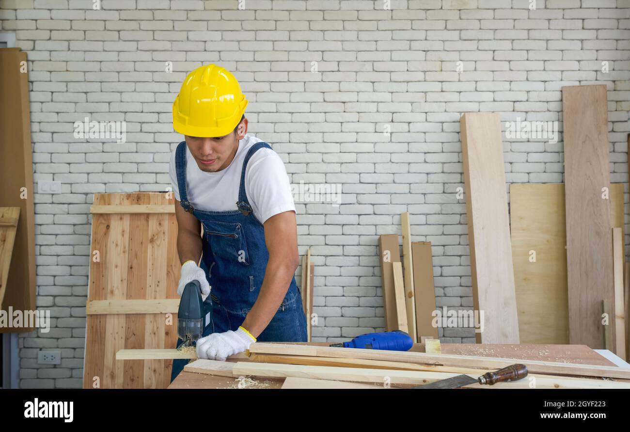 Der asiatische Zimmermann trägt einen gelben Harthut und schneidet in der Holzbearbeitungsanlage mit einer elektrischen Handsäge Sperrholz. Ein Schreibtisch mit Handwerkzeugen und Holzpfählen. Stockfoto