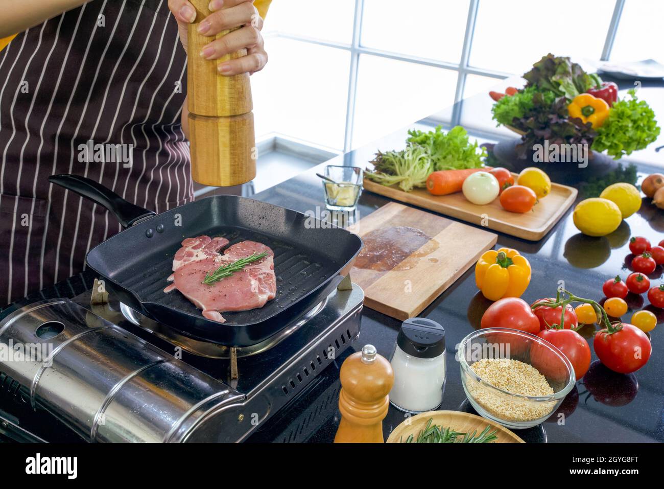 Der Koch setzt Pfeffer auf das Steak. Morgenstimmung in einer modernen Küche. Die Küchentheke voll mit verschiedenen Gemüsesorten und Gewürzen. Stockfoto