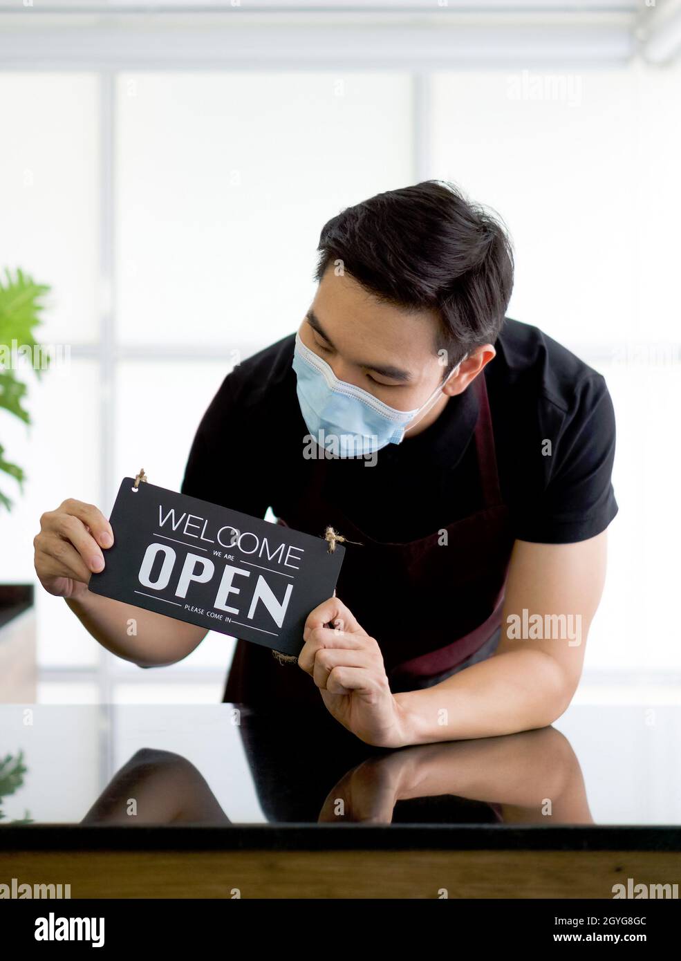 Ein junger asiatischer Mann mit Gesichtsmaske hält ein Eröffnungsschild vor einem Café-Schalter. Stockfoto