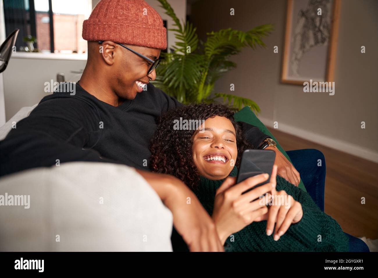 Ein multiethnisches Paar lächelt, während es auf das Mobiltelefon schaut. Moderne Wohnung auf dem Sofa Stockfoto