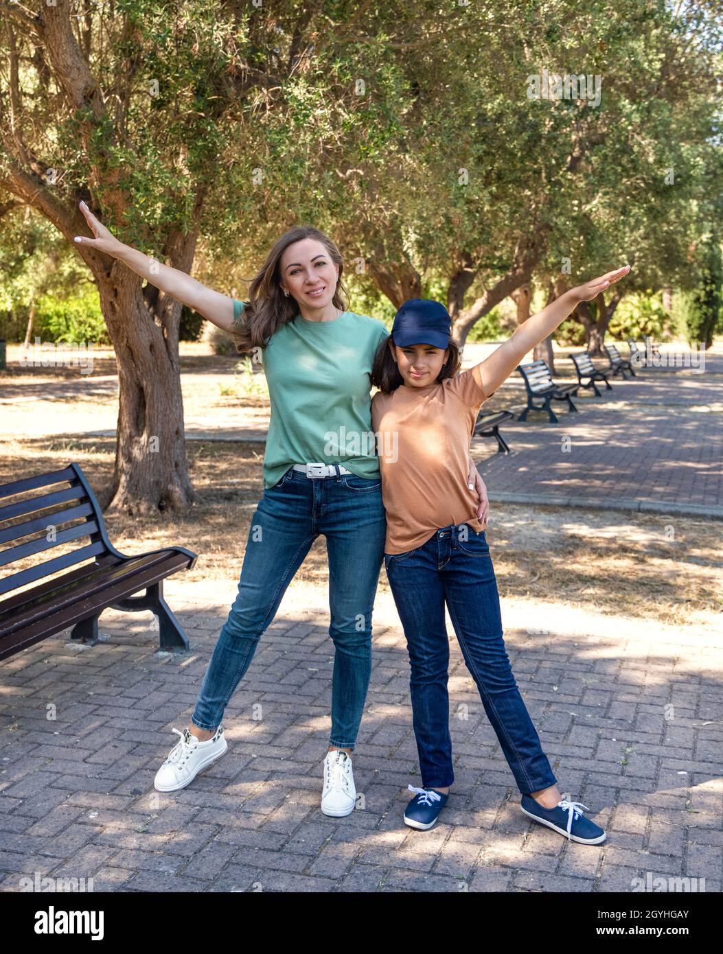 Mutter mittleren Alters und ihre kleine Tochter stehen im Park draußen und schauen auf die Kamera. Liebevolle Familie trägt T-Shirts und Jeans und verbringt Zeit zusammen Stockfoto