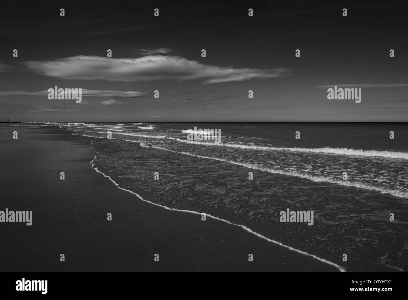 Schwarz-weiß-Foto mit dunklem Thema vom Sandstrand mit sanften Wellen, die brechen und hellen Wolken über dem Horizont. Mundesley Beach, Norfolk, England. Stockfoto