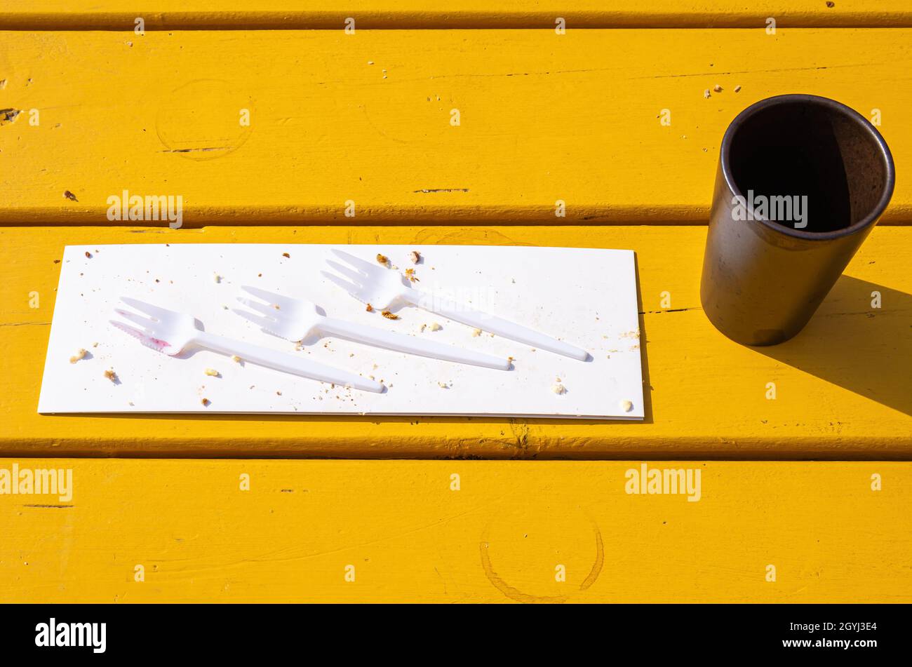 Drei Kunststoffgabeln auf einem Stück Pappe mit Kuchenbröseln und einem leeren Kaffeebecher auf einem gelben Holztisch. Stockfoto