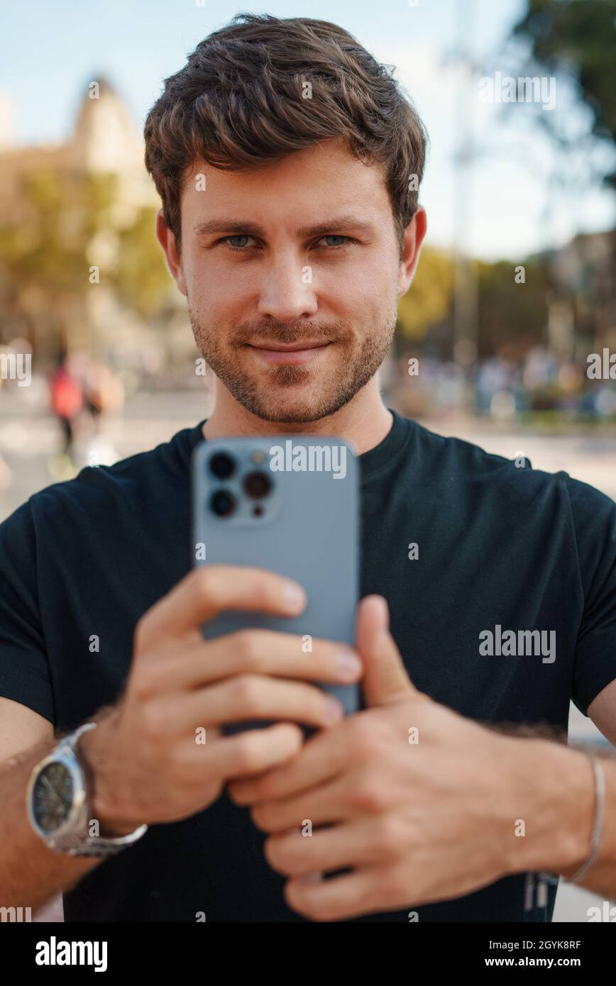 Selbstbewusster junger unrasierter Mann in legerer Kleidung, der am sonnigen Tag auf dem Stadtplatz auf dem modernen Mobiltelefon fotografiert und die Kamera anschaut Stockfoto