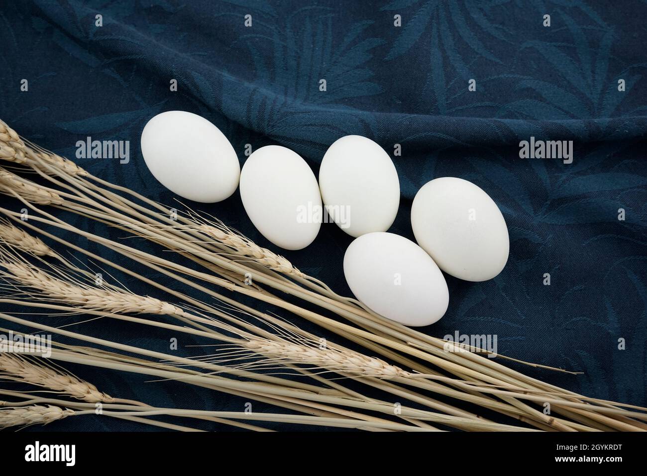 Weiße Eier mit trockenen Weizenstämmen lagen flach auf blauer Charade und grauem Büchsenrauch gefärbtem Bio-Teetuch mit zerknitterter Textur, Serviette, Stoff abstrakt. Stockfoto