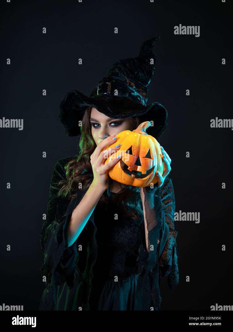 Schöne Hexe in einem spitzen Hut hält einen Kürbis mit einem lachenden Gesicht, Jack Laterne. Halloween-Party, Hexen Sabbat. Junge schöne Brünette in Stockfoto