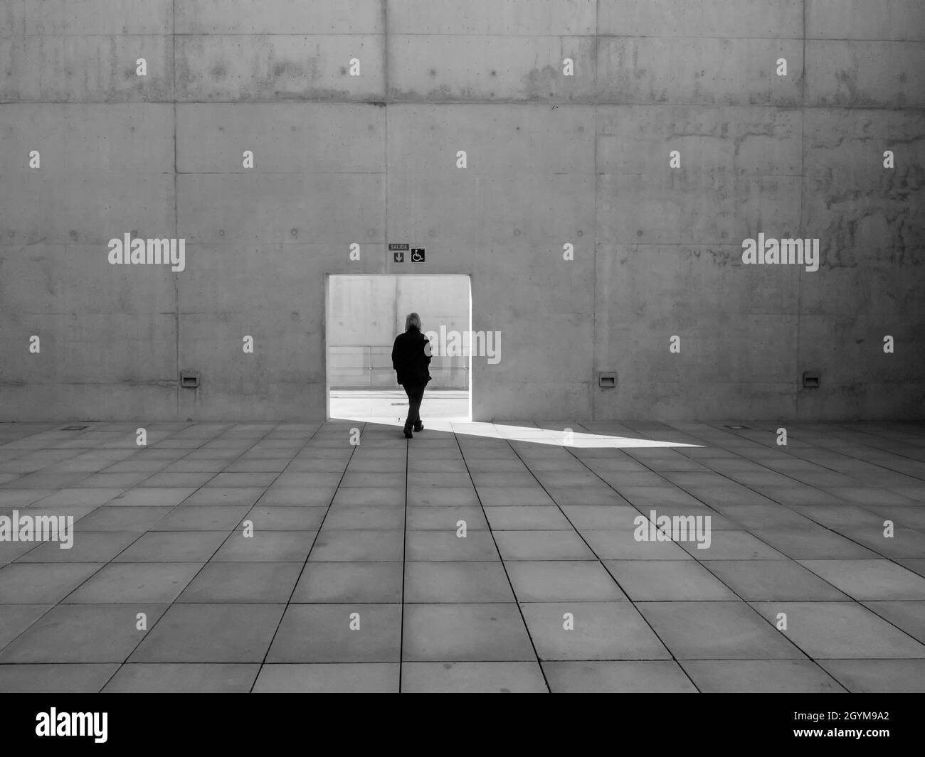 Frau allein, die durch eine quadratische Tür in einer Betonwand herauskommt Stockfoto