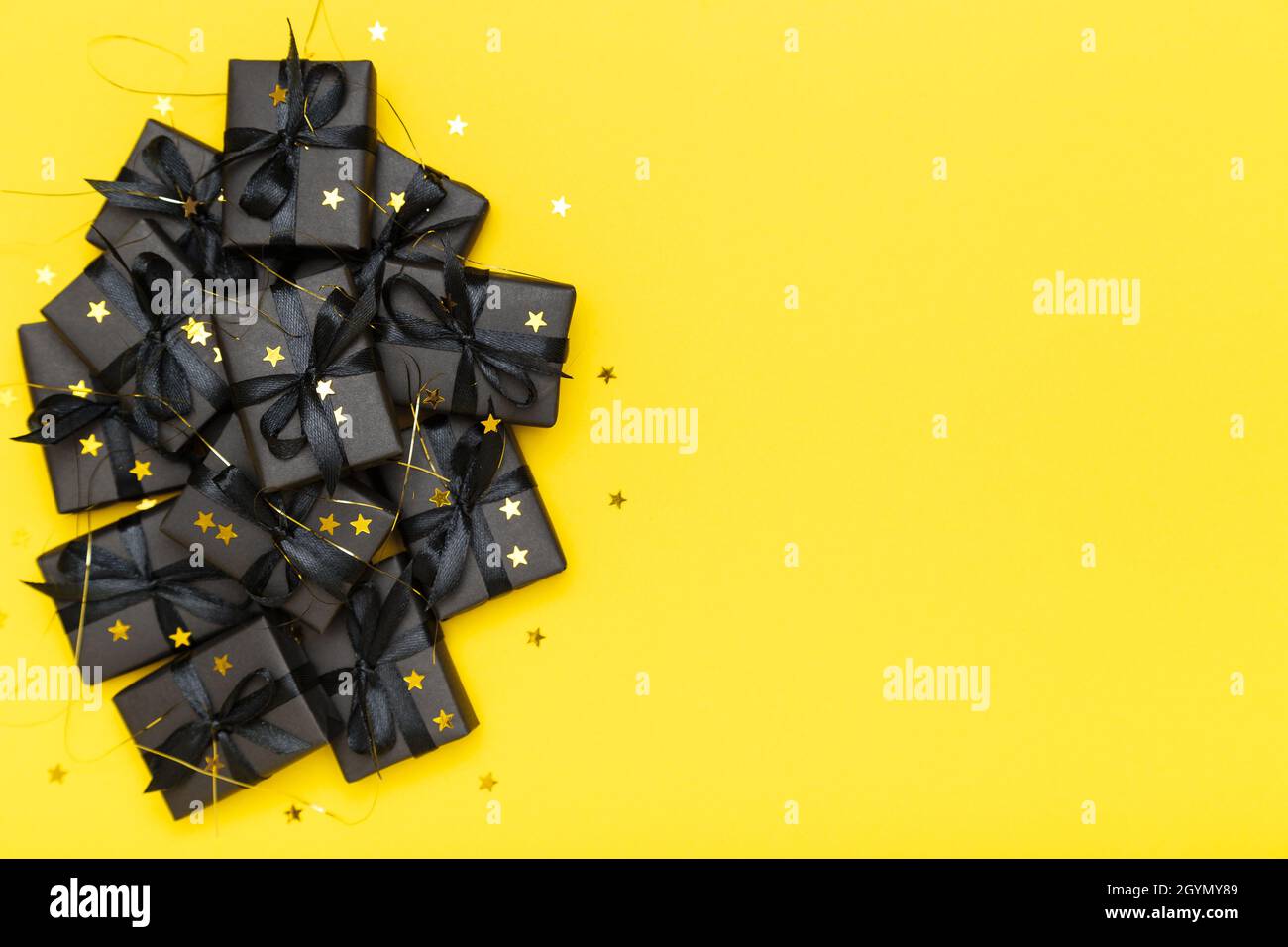 Schwarze Geschenkschachteln mit schwarzen Bändern und goldenen Glitzersternen-Konfetti funkeln auf hellem, sattem, gelbem Hintergrund. Black Friday Sale flat lay, Draufsicht, Kopierfläche Stockfoto
