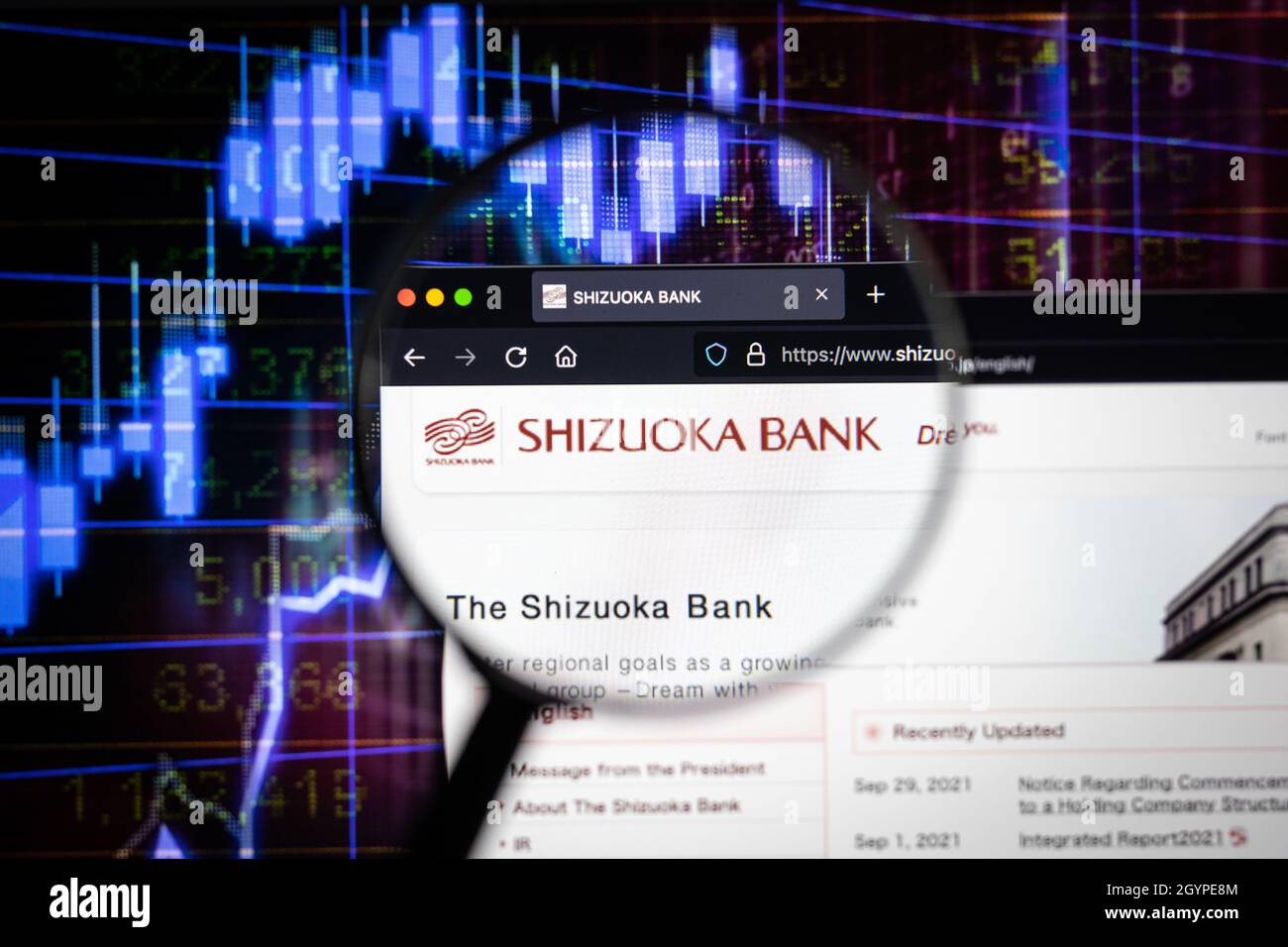 Firmenlogo der Shizuoka Bank auf einer Website mit verschwommenen Börsengrafiken im Hintergrund, die auf einem Computerbildschirm durch eine Lupe gesehen werden. Stockfoto