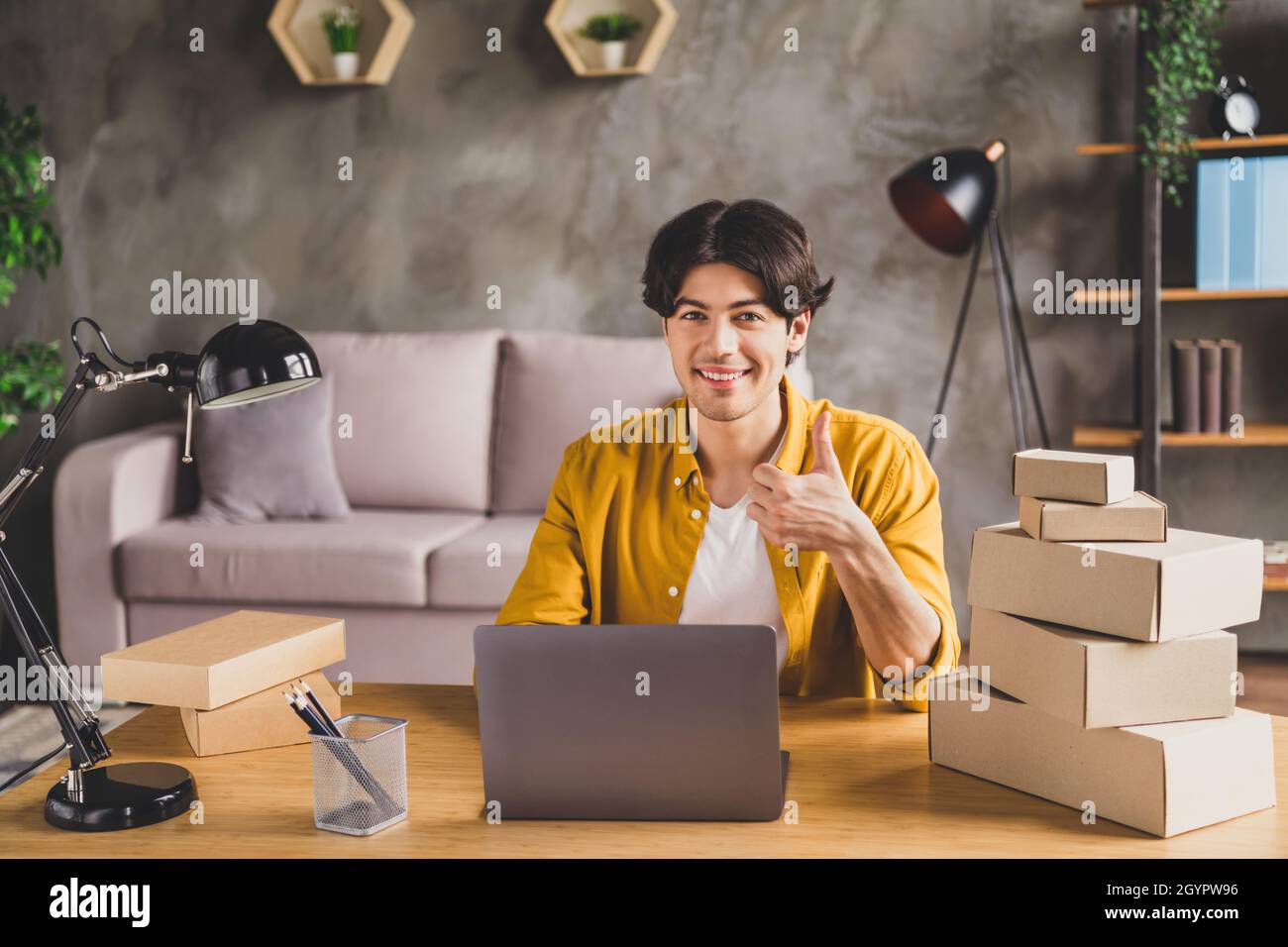 Foto von zuverlässigen Promoter Kerl sitzen Desktop Heben Daumen hoch Netbook Stapel Karton-Box tragen gelbe Hemd nach Hause drinnen Stockfoto