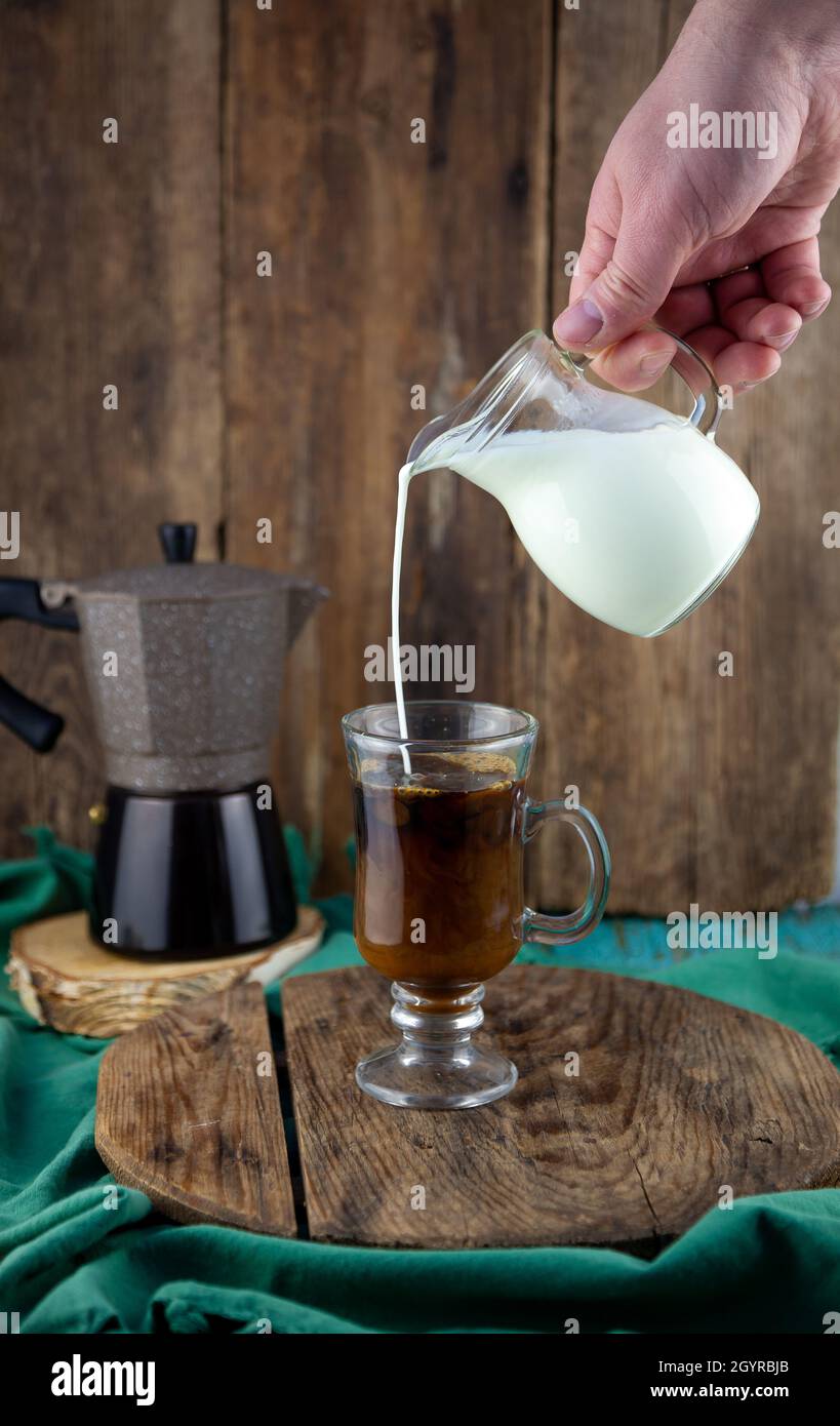 Trinken Sie in einer Tasse. Die Milch wird in den Dekanterkaffee gegossen. Geyser Kaffeemaschine, Milchpulver auf einem Holztisch. Tasse aus Glas mit Cappuccino. Kaffee zubereiten, Hand aus nächster Nähe. Stockfoto