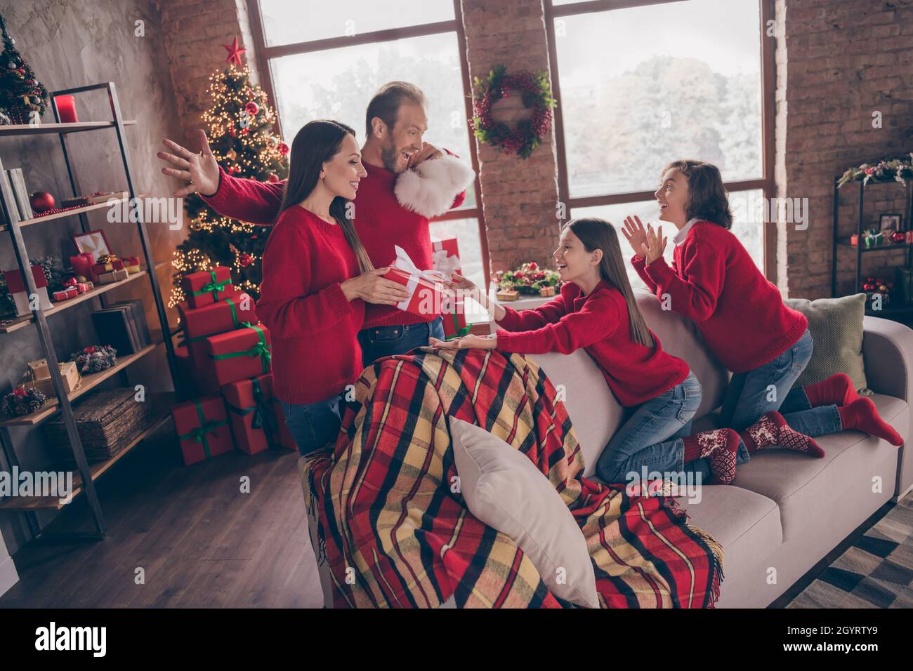 In voller Länge Profilfoto von aufgeregt Familie Bruder Schwester nehmen Geschenke sitzen auf Couch tragen rote Pullover Jeans Socken drinnen zu Weihnachten Stockfoto