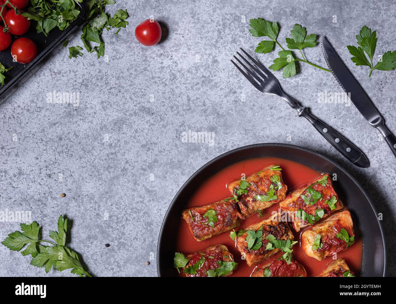 Kohlröllchen in Tomatensauce und gehackter Petersilie in der Pfanne. Flach mit Kohlrollen, einigen Zutaten, Gabel und Messer auf grauem Stein-Bac legen Stockfoto