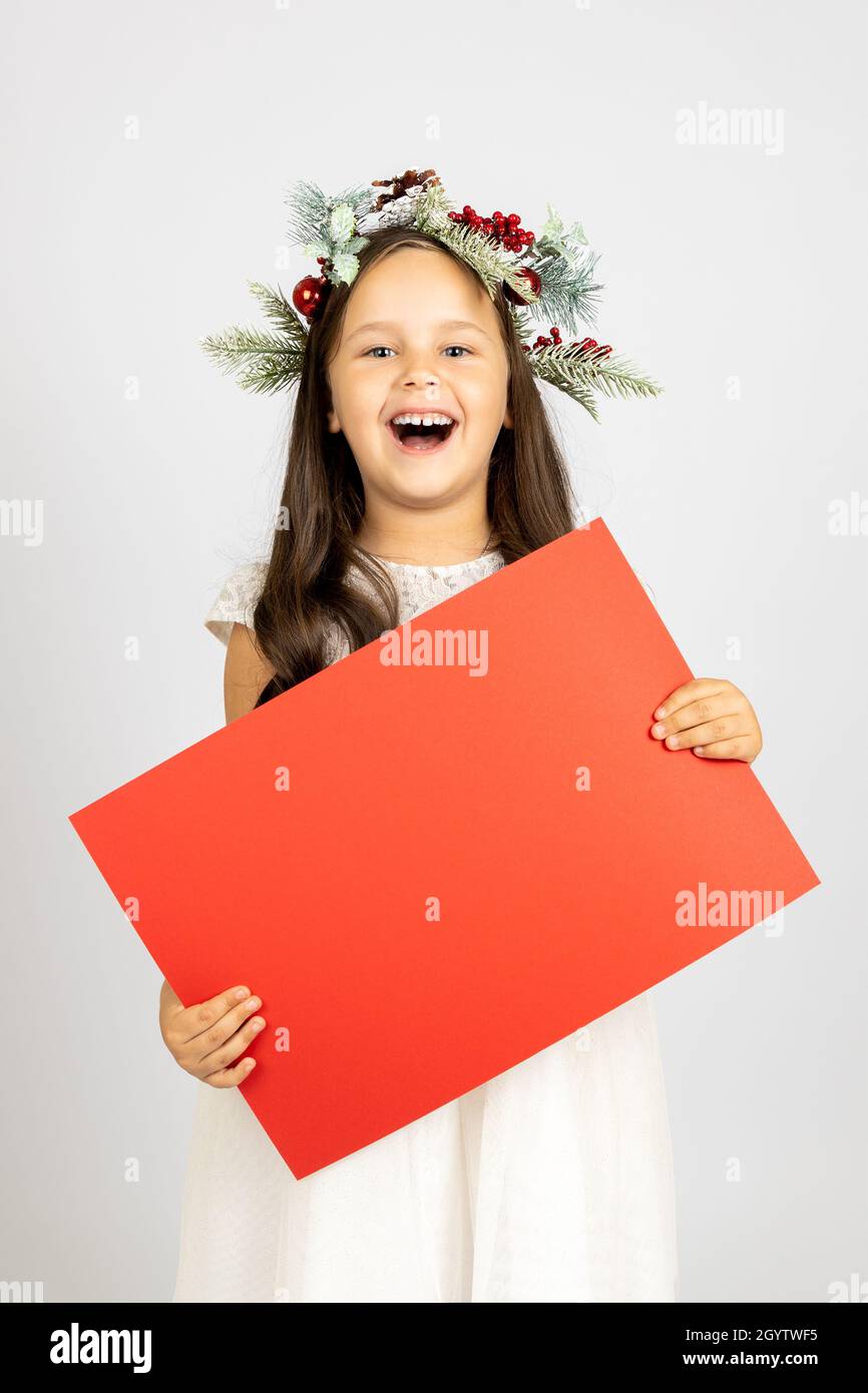 Überglücklich, glückliche Mädchen in einem Weihnachtskranz, die ein rotes, leeres Papier mit einem isolierten Kopierraum auf weißem Hintergrund halten Stockfoto