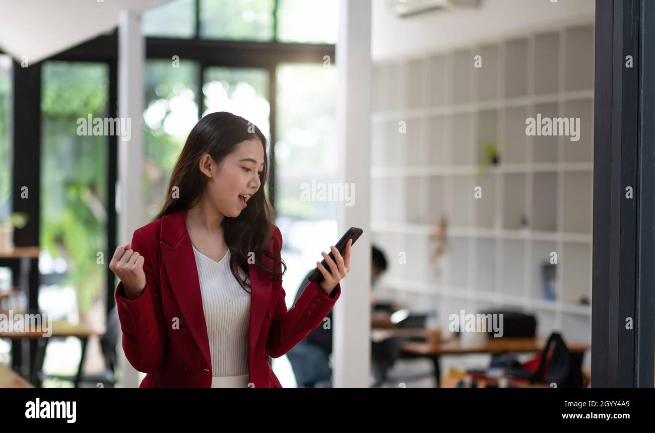Lächelnde asiatische Frau, die das Mobiltelefon mit der Faust hält und für den Erfolg im Büro begeistert ist Stockfoto