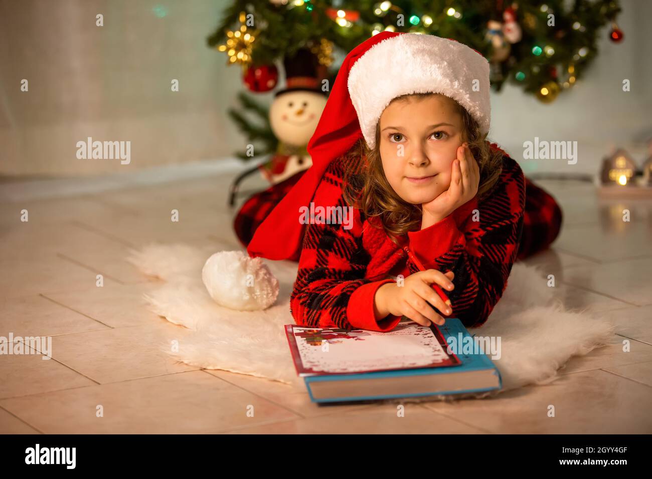 Ein Mädchen liegt auf dem Boden, träumt und schreibt einen Brief an den Weihnachtsmann, im Hintergrund steht ein festlicher Weihnachtsbaum. Ein Kind in einem Hut von Santa Stockfoto