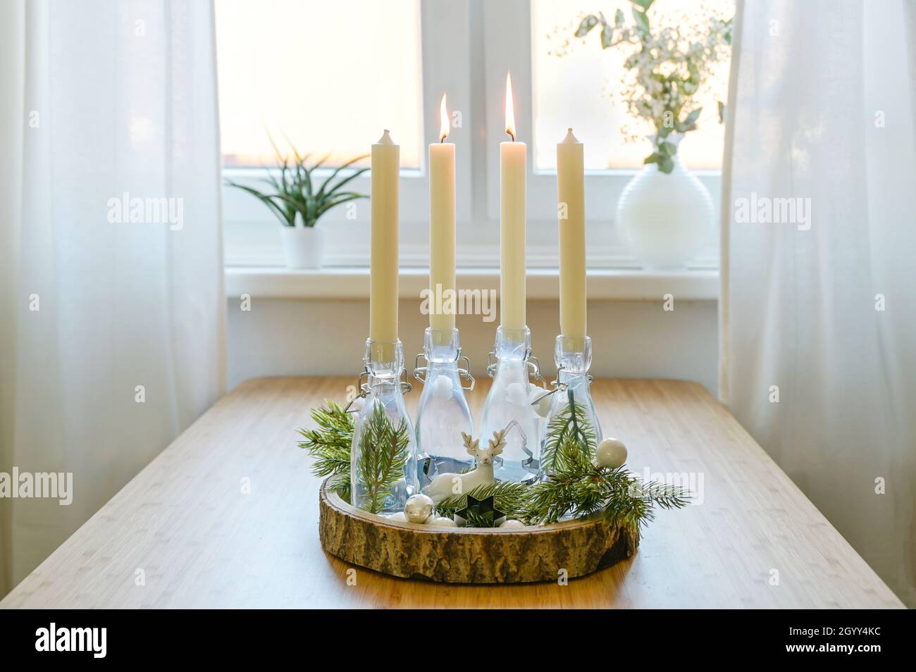 Alternativer Adventskranz, Kerzen in Flaschen auf einem Holzbrett mit Dekoration auf einem Tisch am Fenster, zwei werden angezündet, zweiter sonntag vor weihnachten, Stockfoto