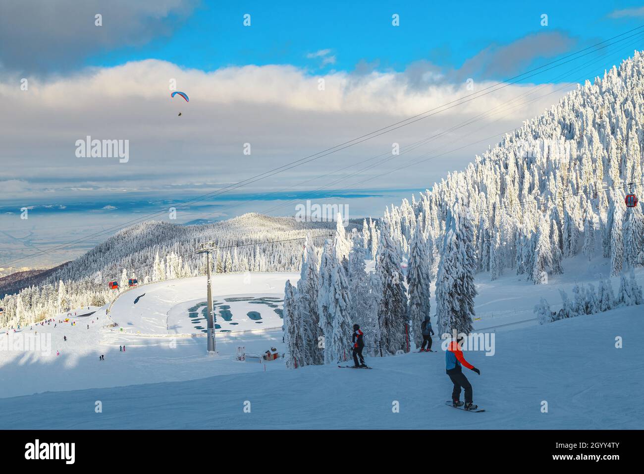 Sportliche Skifahrer und Snowboarder auf der Skipiste im schneebedeckten Kiefernwald. Paragliding über dem Skigebiet, Poiana Brasov, Karpaten, Rumänien, Stockfoto