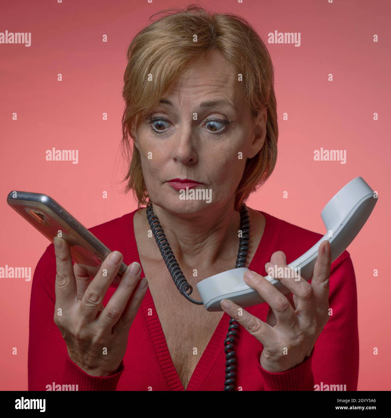 Eine weiße Frau, die in Rot auf einem roten Hintergrund gekleidet ist, hält zwei Telefone in ihren Händen und weiß nicht, welches sie beantworten soll, mit einem lustigen Ausdruck Stockfoto