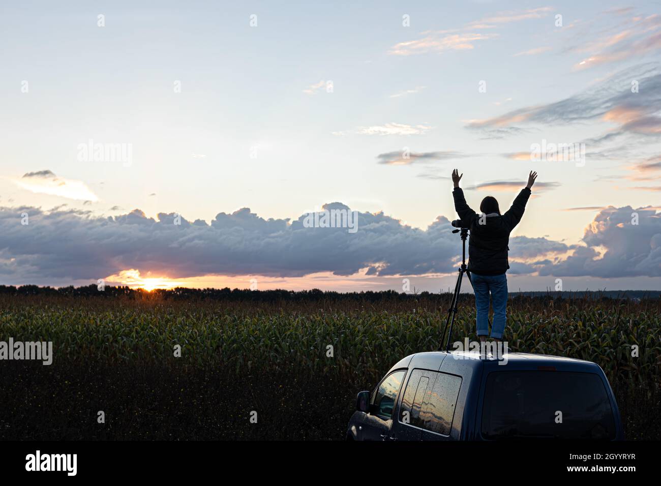 Ein Mädchen fotografiert den Sonnenuntergang von einem Stativ aus, das auf dem Dach eines Autos auf dem Feld steht. Stockfoto
