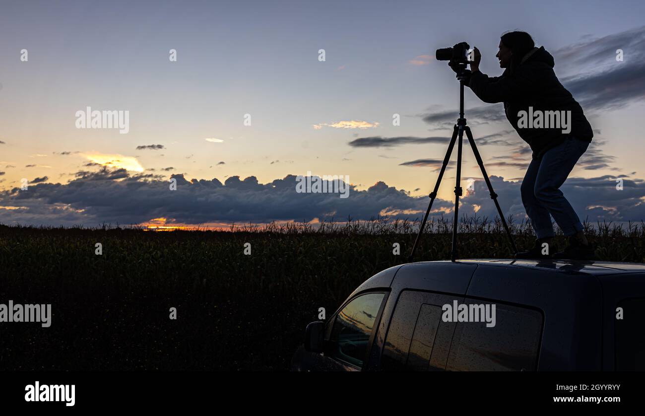 Ein Mädchen fotografiert den Sonnenuntergang von einem Stativ aus, das auf dem Dach eines Autos auf dem Feld steht und den Raum kopiert. Stockfoto