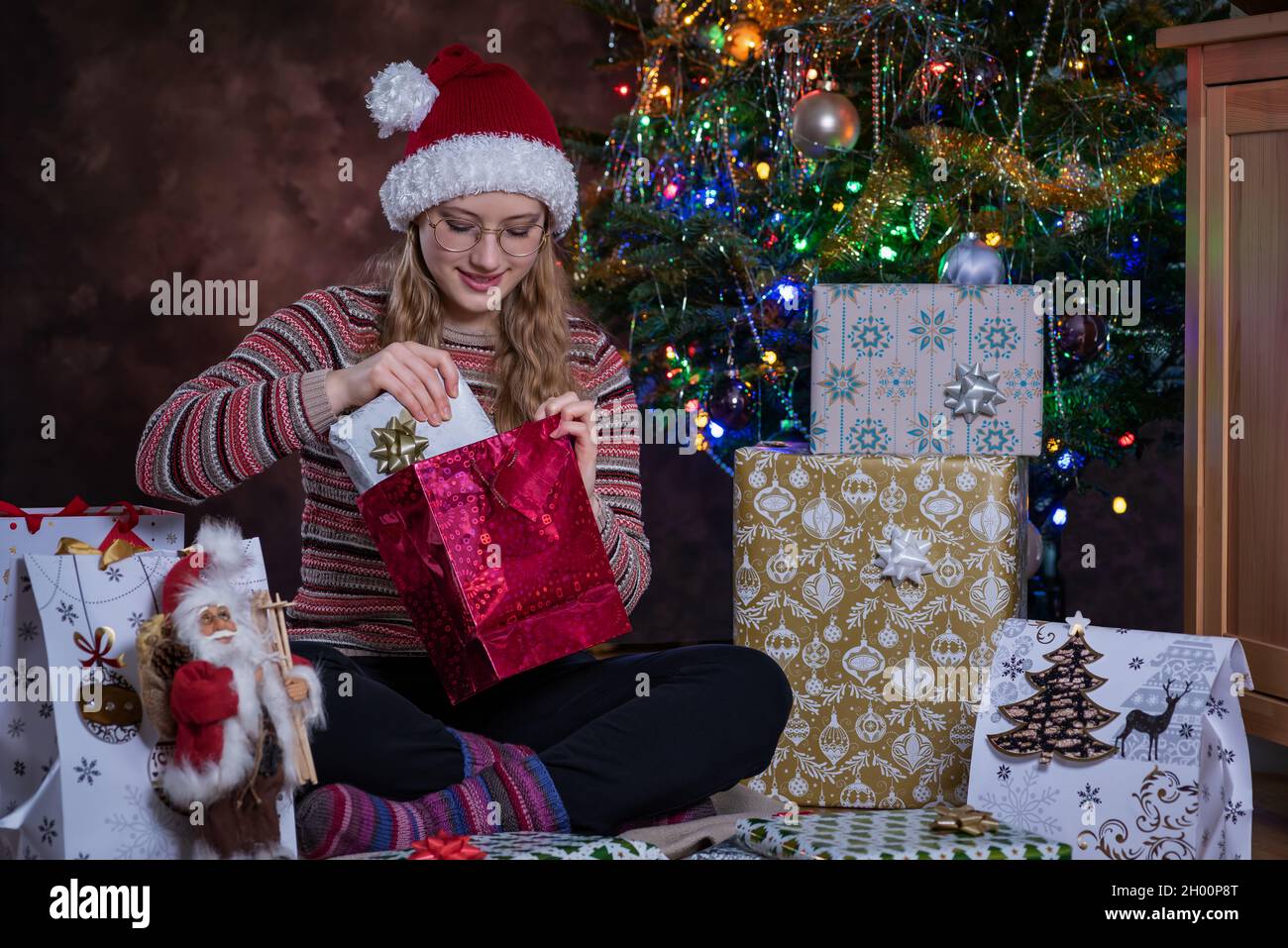 Ein Mädchen, das eine Brille und einen Weihnachtsmann-Hut trägt, lächelt mit einer fröhlichen Stimmung, sitzt neben einem Weihnachtsbaum und öffnet Geschenke. Weihnachtsgeschenke. Überraschung. Stockfoto