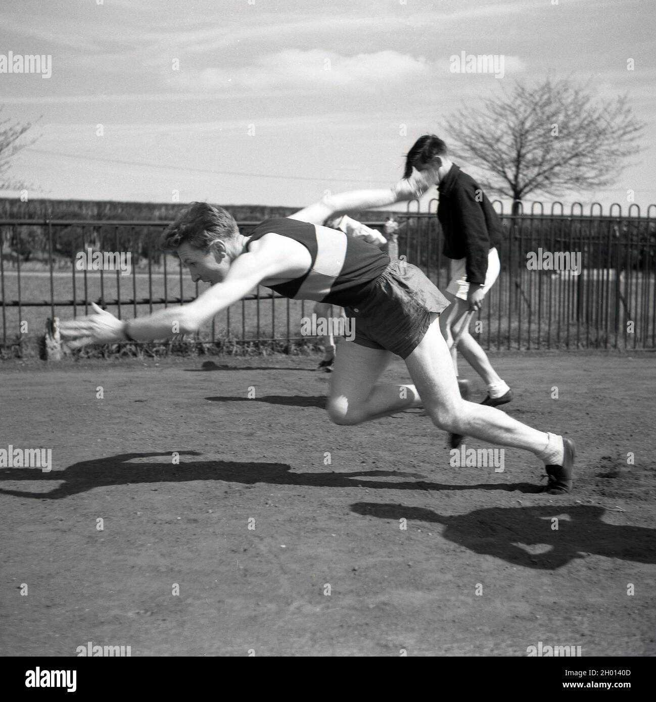 1960, historisch, draußen auf einem Seitengrasgelände an einem Zaun auf einem Sportplatz, ein junger erwachsener männlicher Athlet in Weste, kurz- und Sportschuhen, möglicherweise ein Sprinter, in einer hockigen Position mit den Armen aus, übt seinen Start, England, Großbritannien. Stockfoto