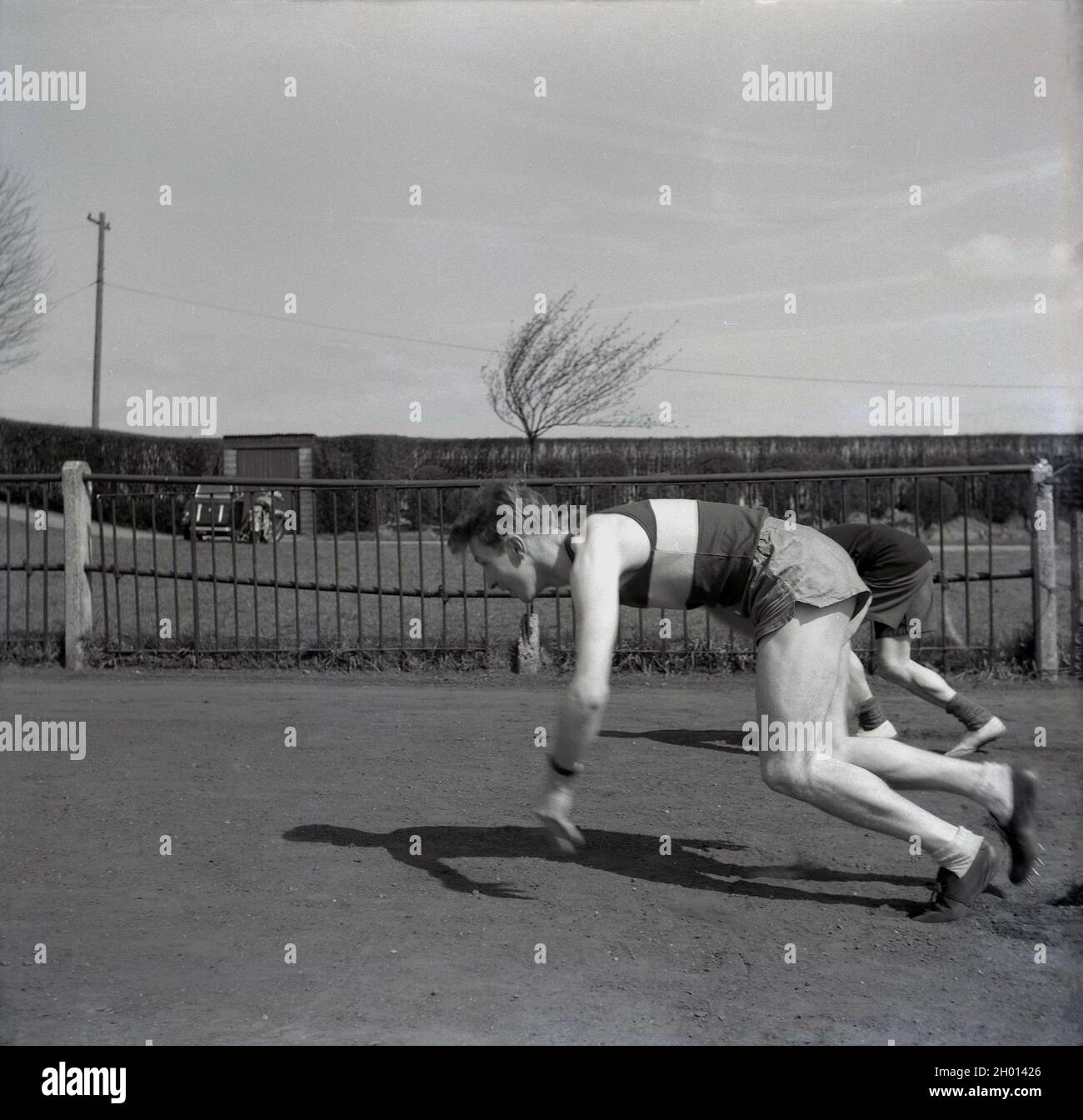 1960, historisch, draußen auf einem Seitengrasgelände an einem Zaun auf einem Sportplatz, ein junger erwachsener männlicher Athlet in Weste, kurz- und Sportschuhen, möglicherweise ein Sprinter, in einer hockigen Position, der seinen Start praktiziert, England, Großbritannien. Stockfoto