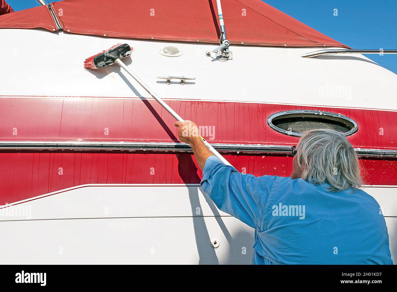 Älterer kaukasischer Mann, der ein rot-weißes Boot mit einer Bürste mit langem Griff wascht Stockfoto