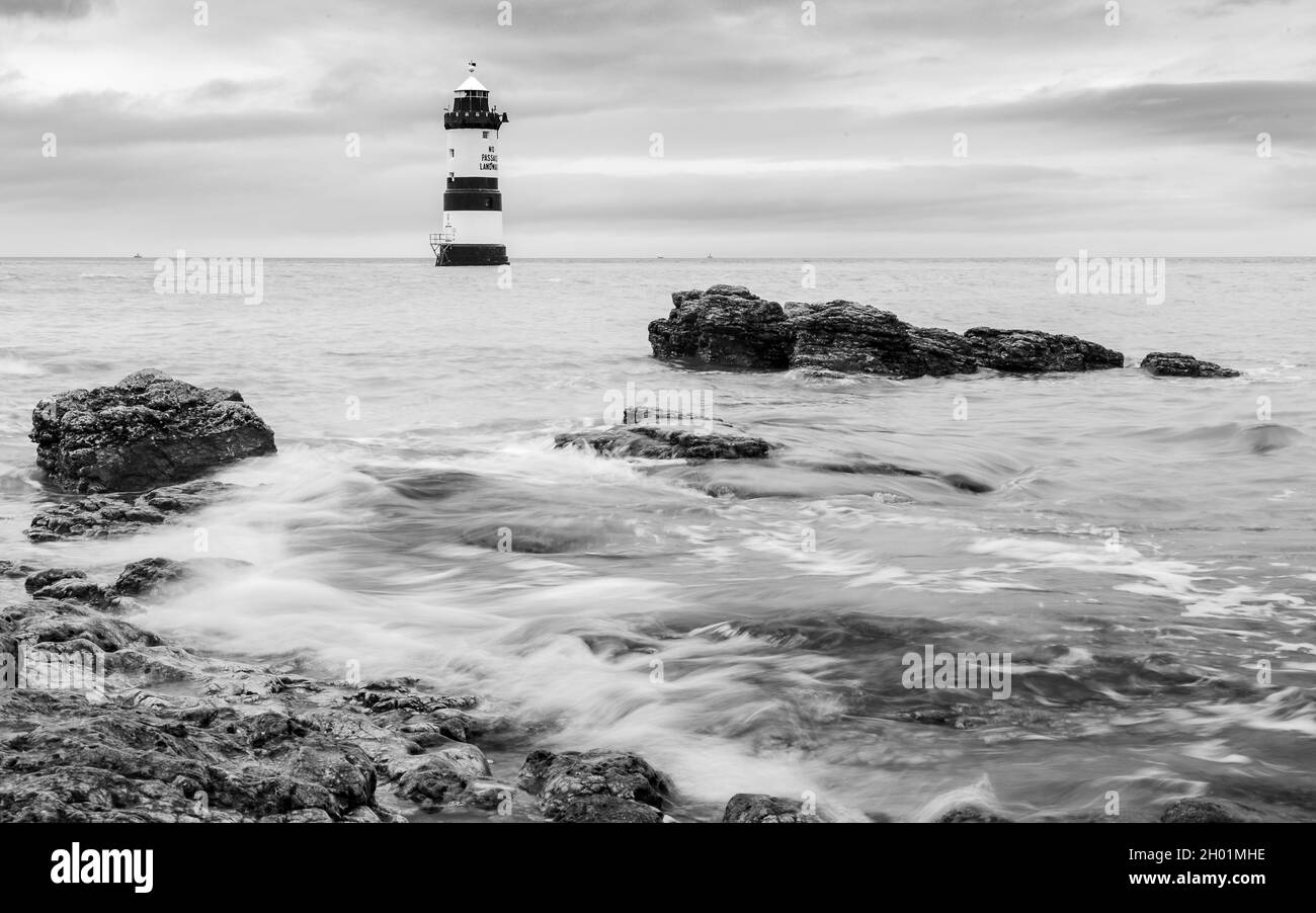 Schwarz-weißes Bild von Wellen, die um den Penmon Lighthouse und die zerklüfteten Felsen im Vordergrund herumschlagen. Auf der Anglesey Küste in Nort Stockfoto