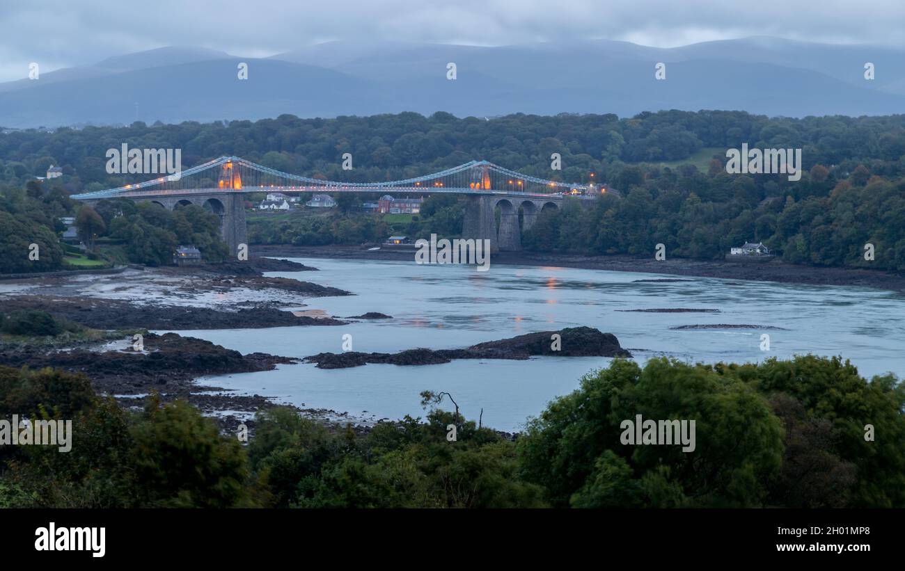 Die Menai Suspension Bridge wurde an einem Abend im Oktober 2021 über die Menai Strait an der Küste von Nordwales hinweg gesehen, als das Licht verblasst. Stockfoto