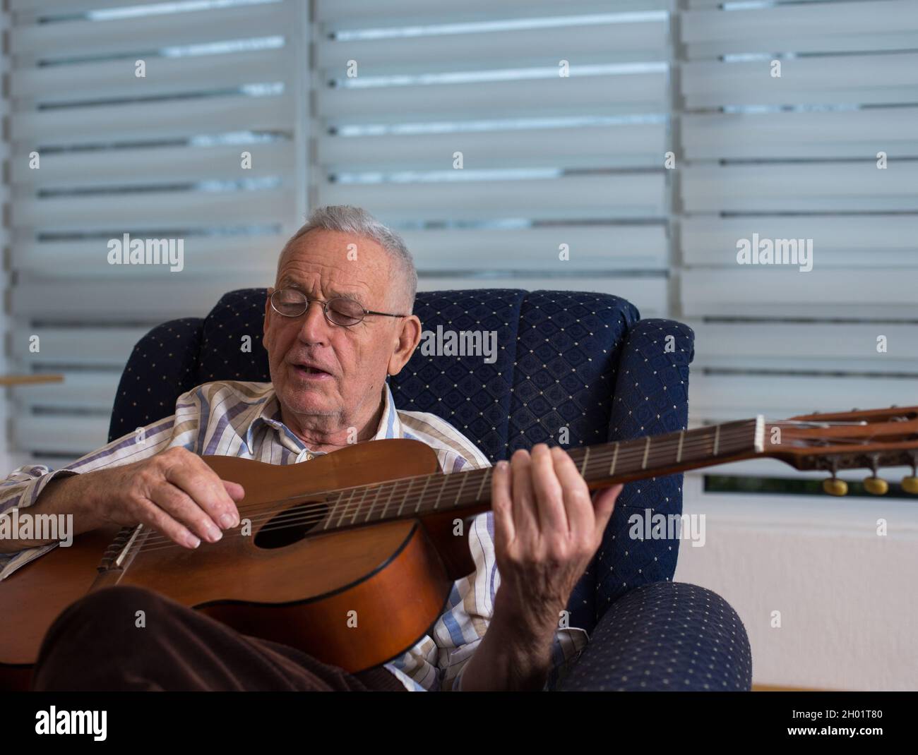 Glücklicher alter Mann, der zu Hause Akustikgitarre spielt und in seinem Sessel singt Stockfoto