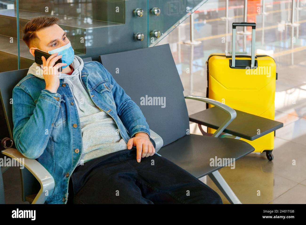 Porträt eines attraktiven jungen Mannes, der vor der Reise mit dem Flugzeug in der Flughafenlounge mit dem Koffer auf dem Smartphone spricht. Kaukasischer Mann im schützenden Gesicht Stockfoto