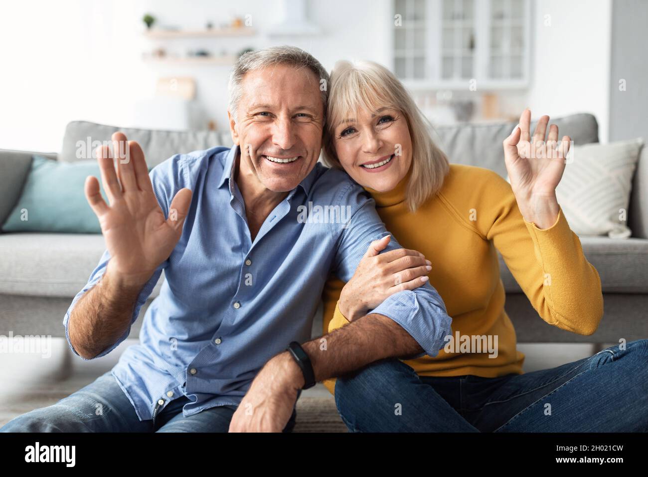 Hallo. Fröhliches Älteres Paar Winkt Die Hände Lächelnd Zur Kamera, Posiert Auf Dem Sofa Und Umarmt Sich Zu Hause. Glückliche Ehe Und Beziehung Im Hohen Alter. Stockfoto