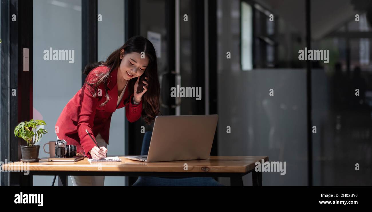 Eine Unternehmerin, die ihr Geschäft vom Büro aus führt, arbeitet an einem Laptop und diskutiert über ihr Geschäft mit dem Smartphone. Stockfoto