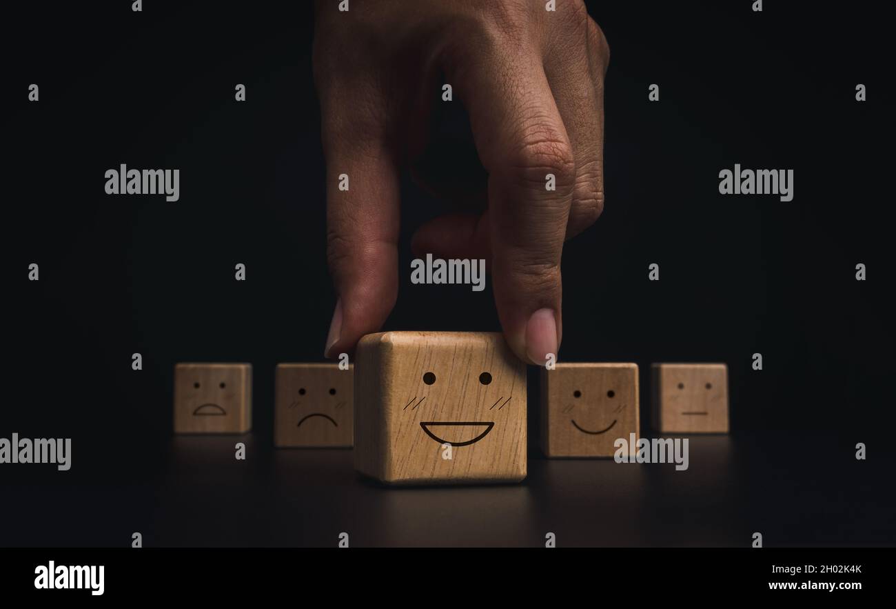Konzept für die Bewertung, Bewertung, Feedback und Zufriedenheitsumfrage durch den Kundenservice. Hand Kommissionierung auf glückliches Lächeln Emoticon Gesicht auf dem Holzblock auf dunkel Stockfoto