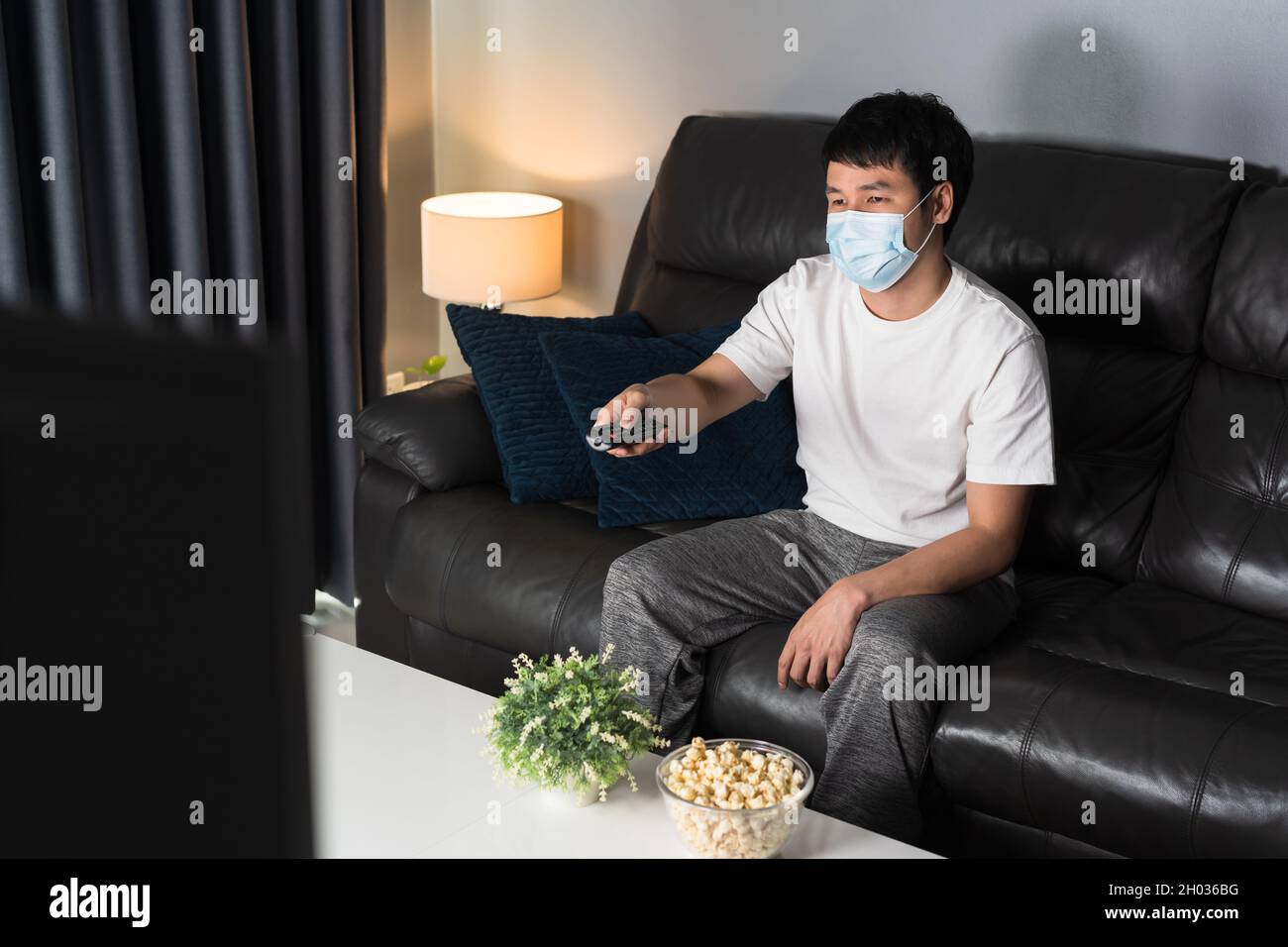 Junger Mann, der auf dem Sofa Fernsehen schaut und eine medizinische Maske trägt, um das Coronavirus zu schützen (Covid-19) Stockfoto