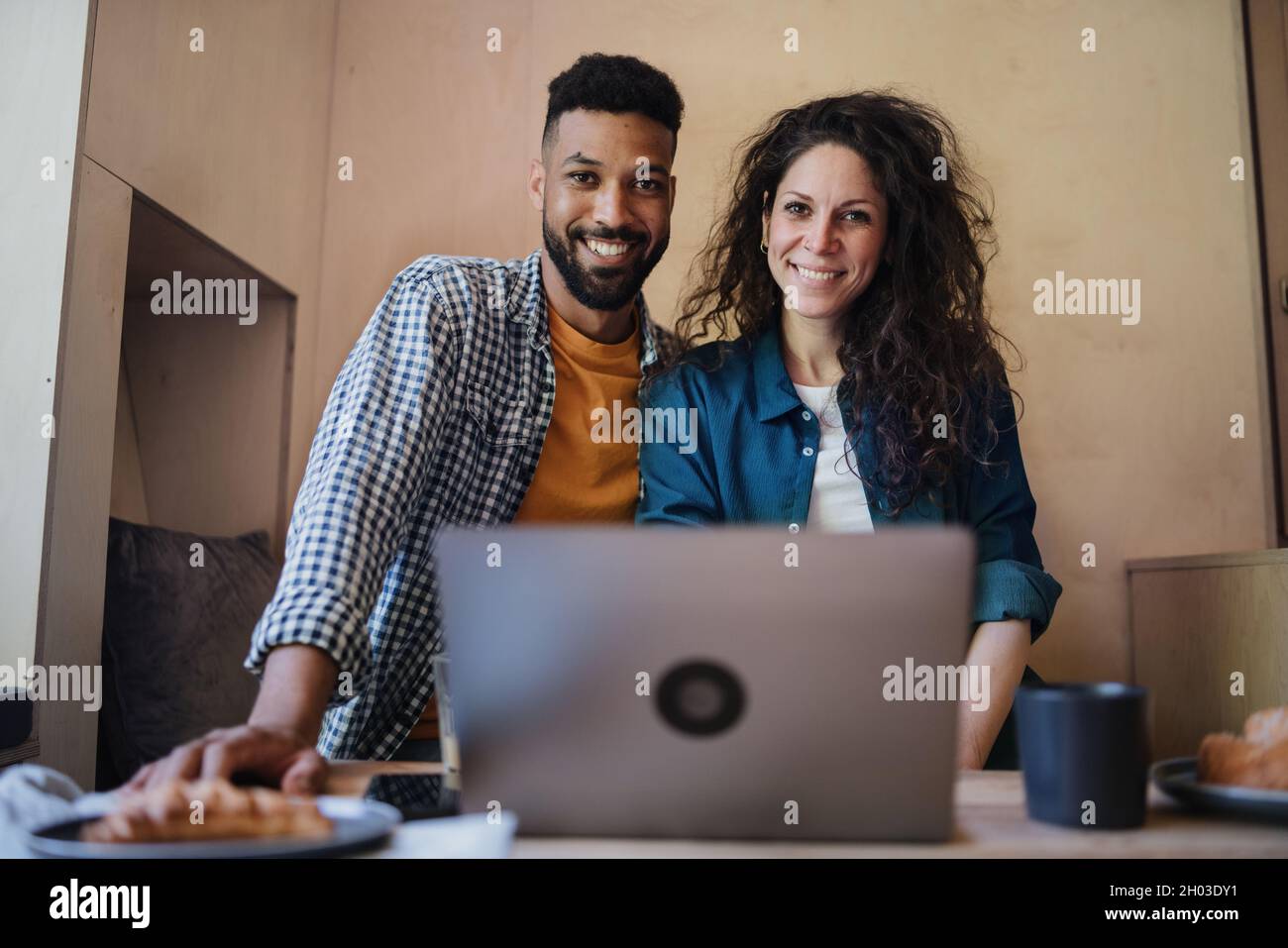 Glückliches biracial Paar mit Laptop sitzen, ruhen und betrachten Kamera drinnen in einem Baumhaus, Wochenende weg und Remote-Office-Konzept. Stockfoto