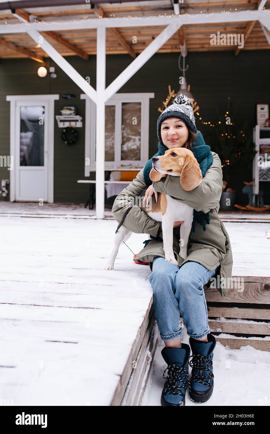 Glückliche Frau, die auf einem verschneiten Terrassenboden vor dem Haus sitzt, mit einem Beagle-Hund auf ihrem Schoß. Lächeln mit einem geschlossenen Lächeln. Stockfoto