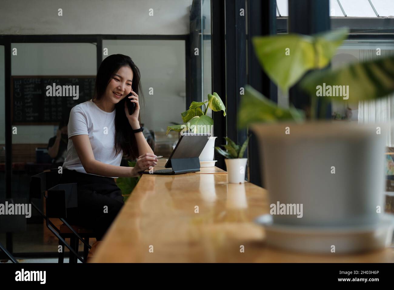 Junge Frau, die auf dem Mobiltelefon spricht und Notizen auf einem digitalen Tablet schreibt, während sie an ihrem Schreibtisch sitzt. Asiatische Frau, die im Heimbüro arbeitet Stockfoto