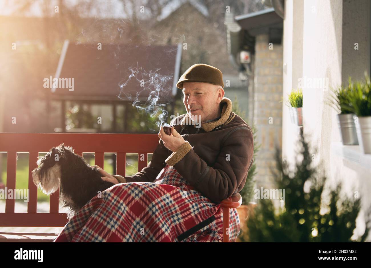 Älterer Mann, der auf einer Bank mit Hund im Garten sitzt, der mit einer Decke und einem Rauchrohr bedeckt ist Stockfoto