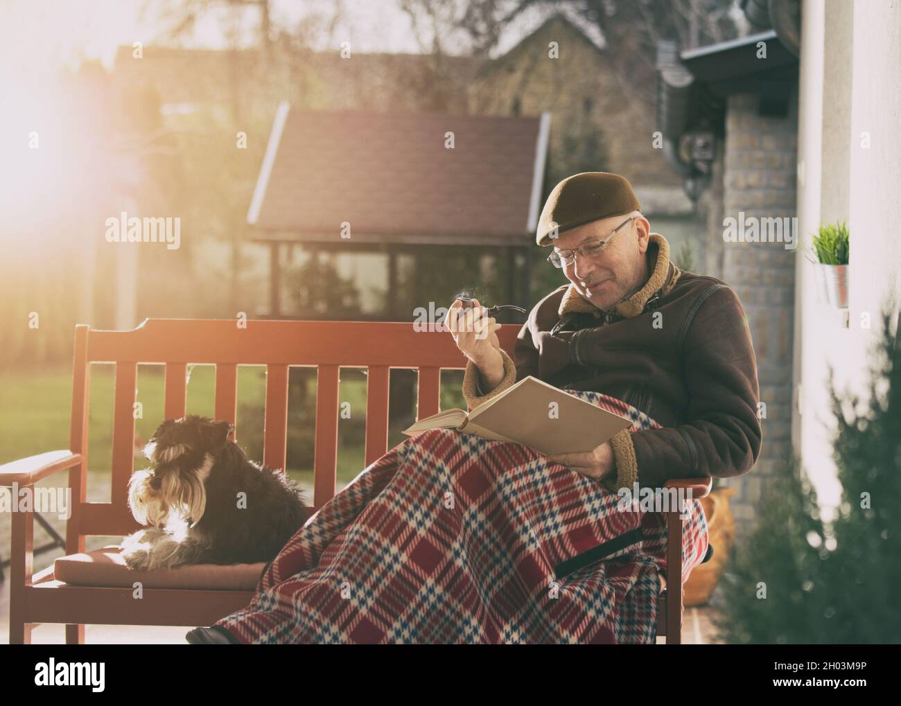 Älterer Mann, der auf einer Bank im Garten sitzt, der mit einer Decke bedeckt ist und ein Buch liest, Pfeife raucht und einen kuscheligen Hund hat Stockfoto