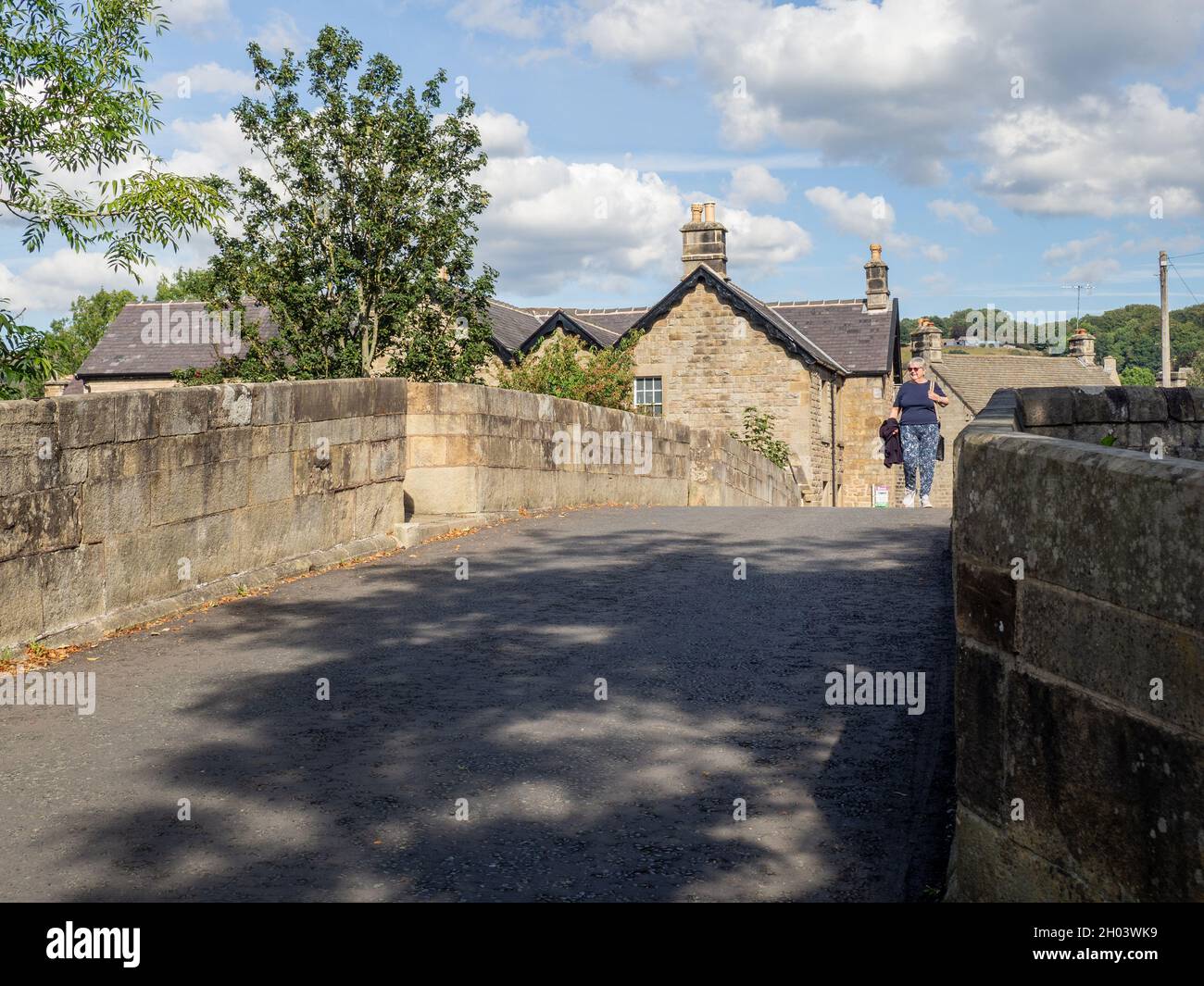 Ältere Frau, die eine alte Brücke aus dem 17. Jahrhundert über den Fluss Derwent im Dorf Baslow, Derbyshire, Großbritannien, überquert Stockfoto