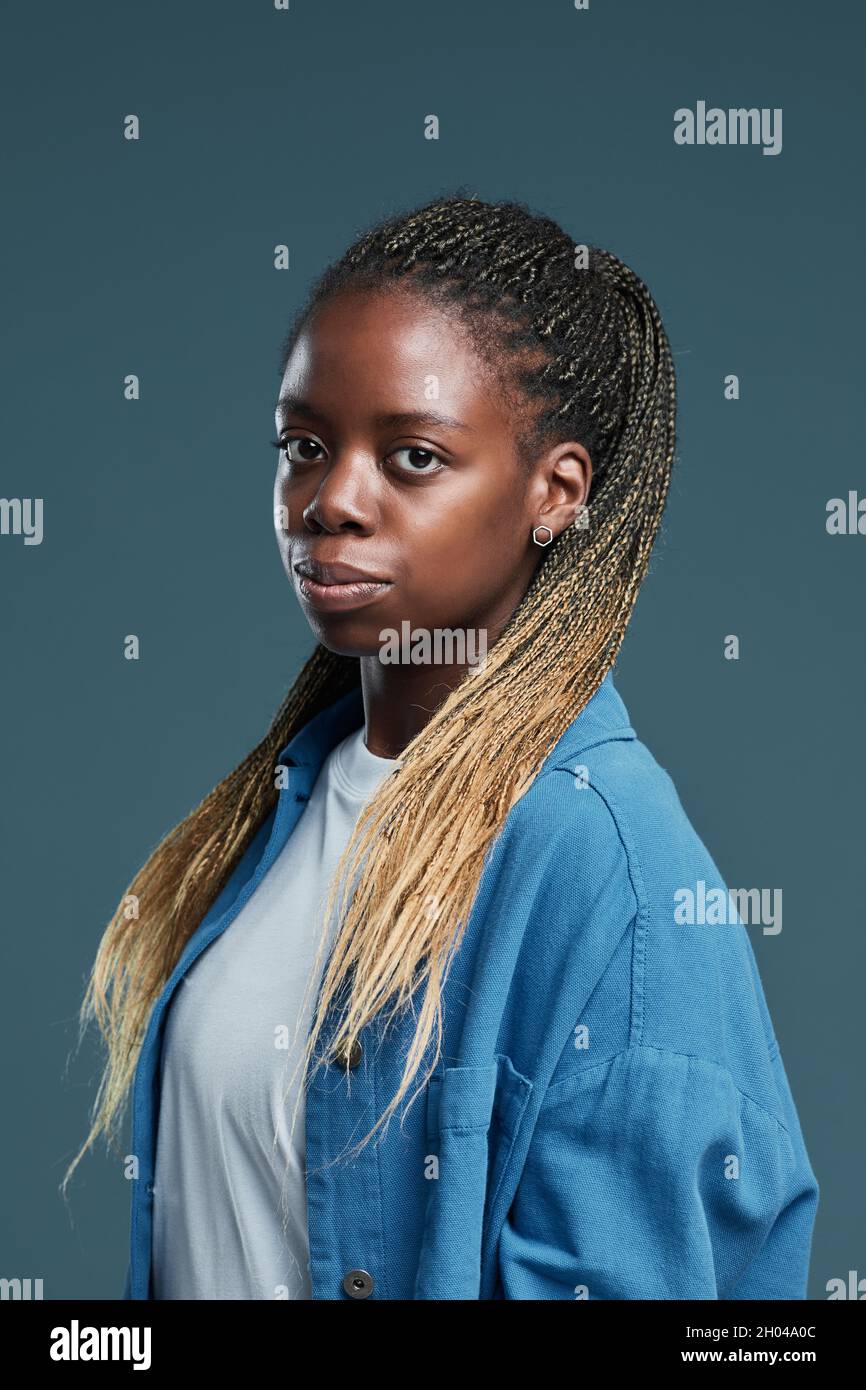 Vertikales Porträt einer modernen afroamerikanischen jungen Frau mit Afro-Zöpfen-Frisur, die vor blauem Hintergrund auf die Kamera blickt Stockfoto