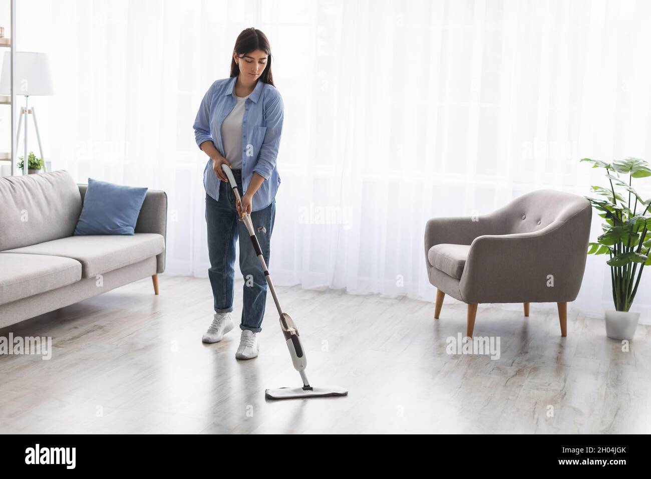 Hausfrau. Casual Frau waschen Hartholz Laminat Boden im Wohnzimmer mit Wasser Spray Mop Pad und nachfüllbare Flasche mit Reinigungsmittel. Verschwommen Stockfoto