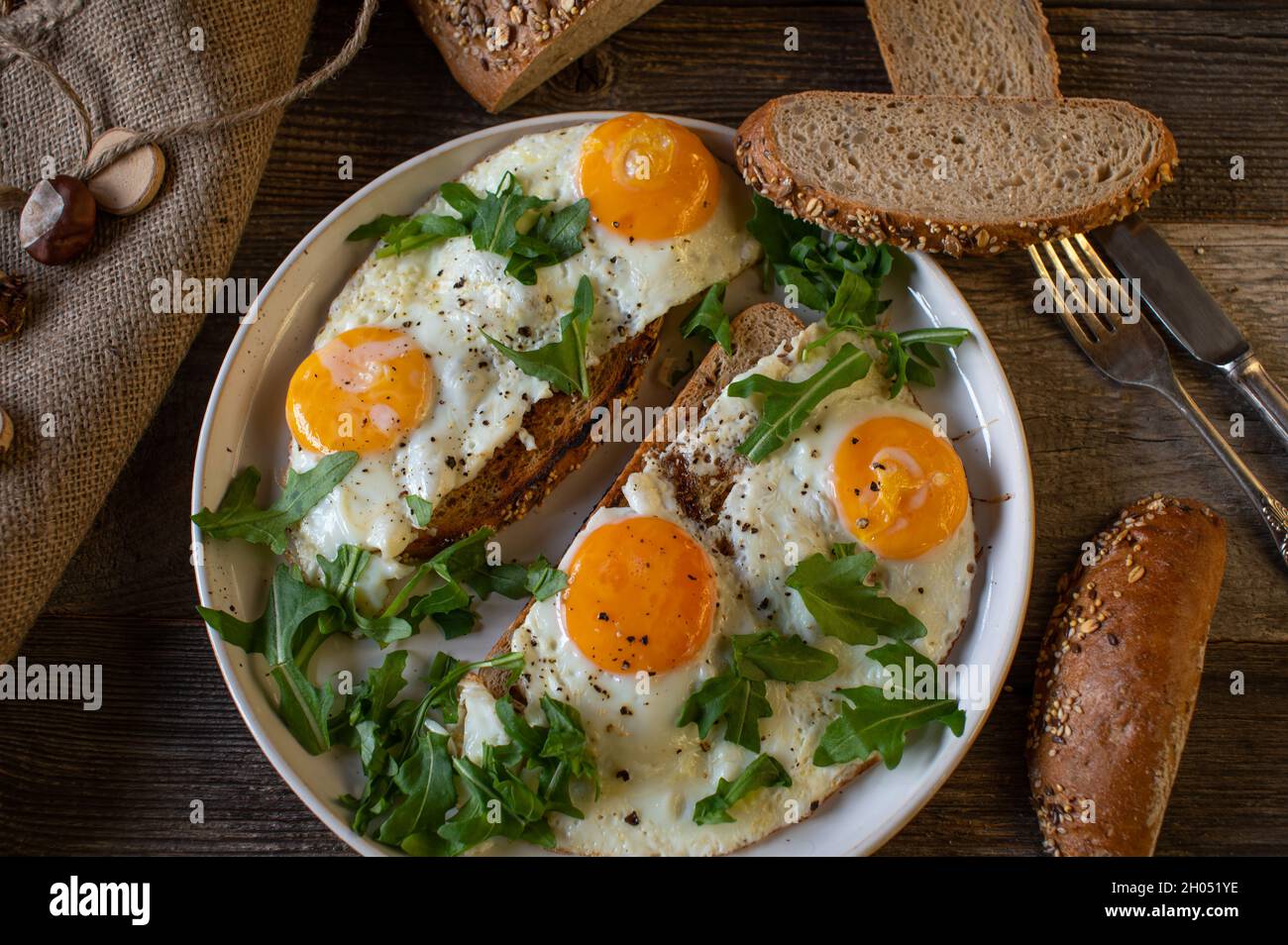 Rustikales Frühstück mit Spiegeleiern, Sonnenseite auf, mit gebratenem Brot und Rucola. Serviert als offenes Sandwich auf einem Teller auf einem Holztisch. Stockfoto