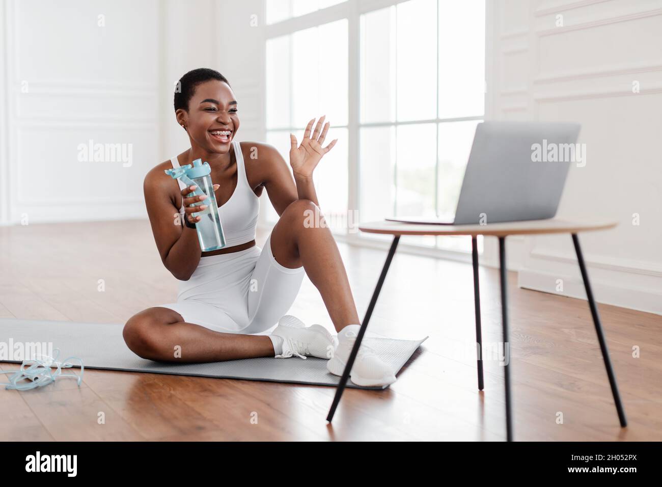 Aufgeregte, sportliche, schwarze Frau in Sportkleidung winkt der Webcam zu, die mit einem persönlichen Trainer oder Followern eine Videokonferenz macht und auf dem Boden Yoga macht Stockfoto