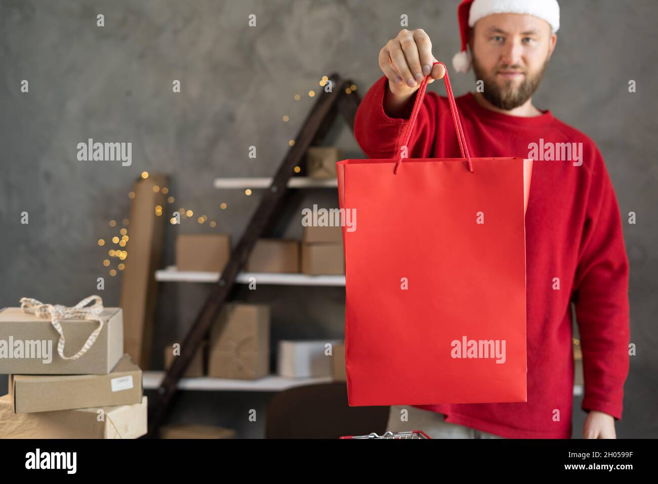 Bärtiger Mann in Weihnachtsmütze mit rotem Weihnachtspaket. Online-Shopping, Lieferung. Inhaber eines Kleinunternehmens in einem Abfertigungslager. Winterangebot. Stockfoto